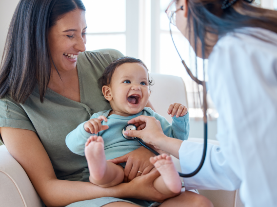 baby laughs on their mother's lap while doctor listens with stethoscope